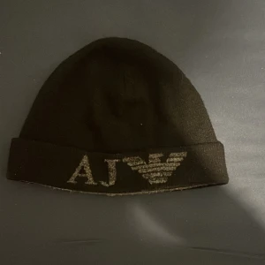Svart mössa från Armani Jeans - Snygg svart mössa från Armani Jeans med AJ-logga framtill. Perfekt för att hålla sig varm och stilren under kyliga dagar. Passar både till vardags och mer uppklädda tillfällen.