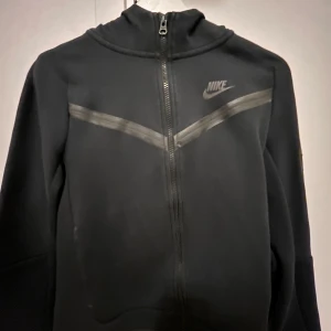Svart Nike teach  från Nike - Säljer en svart hoodie  från Nike i bra skick. Den har en dragkedja framtill och ett diskret märke på bröstet. Perfekt för höst och vår, med en stilren design som passar till det mesta. Jackan är långärmad och gjord i ett mjukt material för extra komfort.