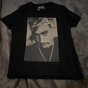 Säljer en svart T-shirt med ett stort tryck av 2PAC på framsidan. T-shirten är i bomull och har en normal passform med korta ärmar. På baksidan finns en text i vitt. Perfekt för alla hiphop-fans! 🖤