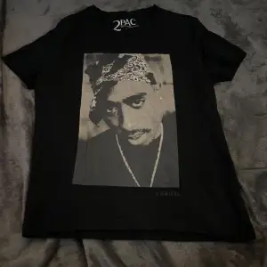 Säljer en svart T-shirt med ett stort tryck av 2PAC på framsidan. T-shirten är i bomull och har en normal passform med korta ärmar. På baksidan finns en text i vitt. Perfekt för alla hiphop-fans! 🖤