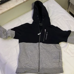 Svart och grå hoodie från Sail Racing - Säljer en snygg hoodie från Sail Racing i svart och grått. Den har en dragkedja framtill och en skön huva. Perfekt för kyliga dagar och passar till både vardag och fritid. Den är i bra skick och redo för nya äventyr!