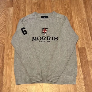Grå stickad tröja från Morris - Säljer en snygg grå stickad tröja från Morris large men passar även Medium. med ett broderat emblem och text på framsidan. Tröjan har långa ärmar och ribbade muddar vid ärmslut och nederkant. Perfekt för höst och vinter!