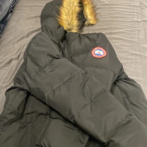 Svart dunjacka från Canada Goose - Säljer en svart dunjacka från Canada Goose med pälsdetalj på huvan. Jackan är perfekt för kalla vinterdagar och har en klassisk design med dragkedja och knappar. Den är i mycket bra skick och håller dig varm och stilren hela säsongen.