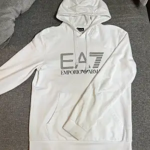 Säljer en snygg vit hoodie från Emporio Armani EA7. Den är i bra skick och har en stor logga på framsidan. Perfekt för en stilren och avslappnad look. Den har en klassisk känguruficka och justerbar huva med dragsko.