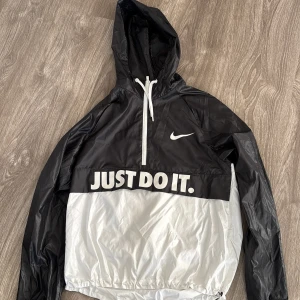 Svart och vit vindjacka från Nike - Säljer en snygg svart och vit vindjacka från Nike med texten 'JUST DO IT.' på framsidan. Jackan har en huva med dragsko och en halv dragkedja framtill. Perfekt för vår och höst när det blåser lite extra. Passar både till träning och vardags.