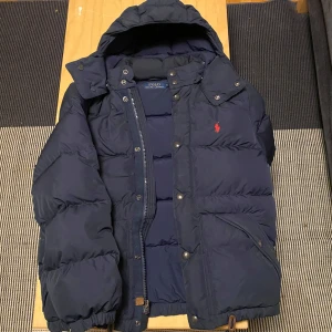 Mörkblå dunjacka från Polo Ralph Lauren - Säljer en snygg mörkblå dunjacka från Polo Ralph Lauren. Jackan har en praktisk huva och stängs med både dragkedja och knappar. Den är perfekt för kalla vinterdagar. Jackan är i bra skick bortom att den har tappat ytterst lite färg på metall knapparna, men det är inget som man lägger märket till. Fortfarande lika puffig som ny, storlek S (sitter lite större)