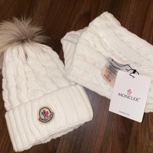 Moncler mössa - Säljer en superfin vit stickad mössa från Moncler med en fluffig pälsboll på toppen. Den har ett snyggt Moncler-märke framtill och är perfekt för kalla vinterdagar. Mössan är i nytt skick och kommer med matchande halsduk. Perfekt för att hålla sig varm och stilren! ❄️