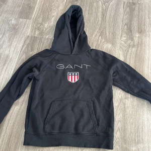 Svart hoodie från GANT - Säljer en snygg svart hoodie från GANT med deras klassiska logga och ett coolt emblem på bröstet. Passar en XS