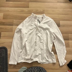 Vit blus/tröja - Säljer en vit blus/tröja med volang detaljer runt halsen samt knappar vid halsen. Aldrig använd