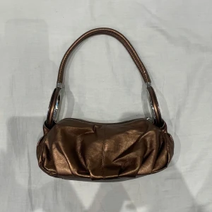 Brun axelväska i fejkskinn - snygg bronsfärgad (brun med lite skimmer i) axelväska i skinn med en stilren design. Väskan har en praktisk dragkedja och en bekväm axelrem. Perfekt för både vardag och fest! Superbra skick och så söt rymmer mobil, lite smink, nycklar typ