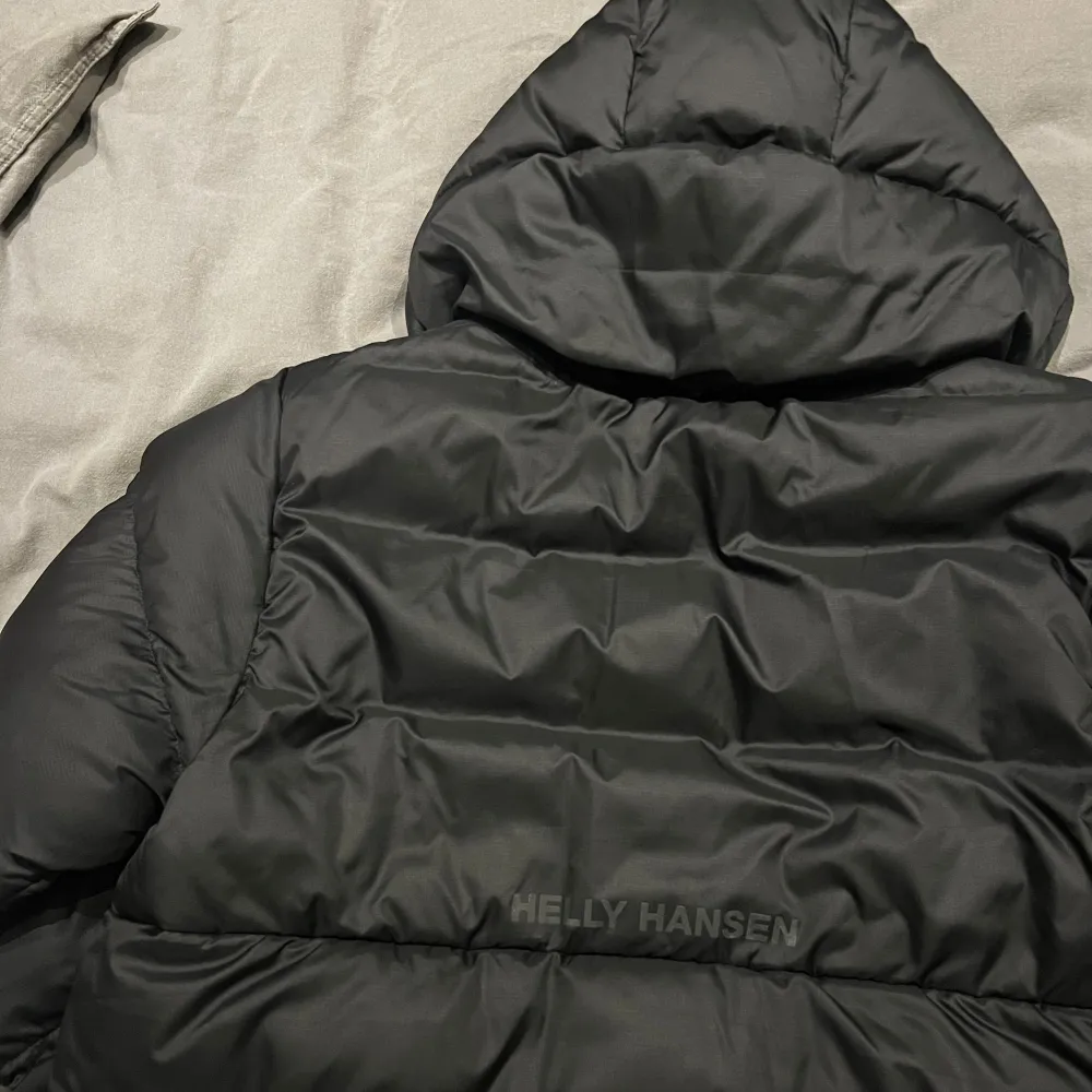 Säljer en svart dunjacka från Helly Hansen i storlek 164. Jackan är perfekt för kalla vinterdagar med sin tjocka vaddering och huva. Den har en stilren design med Helly Hansen-logga på bröstet och ryggen. Jackan är i mycket bra skick och redo för nya äventyr! 🖤Luvan är avtagbar. . Takit.