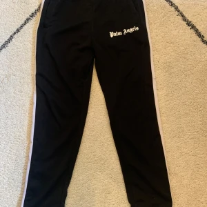 Svarta byxor från Palm Angels - Säljer ett par svarta byxor från Palm Angels med vita sidoremsor. De har en avslappnad passform och är perfekta för både vardag och fest. Byxorna är i mycket bra skick och har en snygg logga på ena benet, för samma 