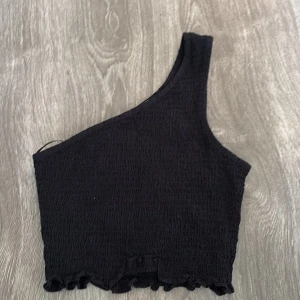 Svart oneshoulder topp - Snygg svart oneshoulder topp i ribbat material. Perfekt för en utekväll eller fest! Den har en cool asymmetrisk design och en liten volangdetalj vid nederkanten. Passar bra till både jeans och kjol.