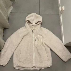 Vit teddyjacka  - Mysig vit teddyjacka med huva, perfekt för kyliga dagar. Jackan har en dragkedja framtill och praktiska fickor. Den är långärmad och har en avslappnad passform. Perfekt för höst och vinter!