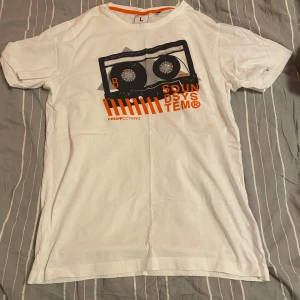Vit t-shirt med tryck från Cropp - Säljer en vit t-shirt från Cropp i storlek L. Den har ett coolt tryck med en kassett och texten 'Sound System' i svart och orange. Perfekt för en avslappnad stil. Regular fit och kortärmad, passar bra till vardags.