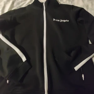 Svart tröja från Palm Angels - Säljer en snygg svart hoodie från Palm Angels i storlek M. Jackan har en vit dragkedja och vita detaljer längs ärmarna. Perfekt för en stilren look, oavsett om du ska på fest eller bara hänga med vänner. Den är i mycket bra skick och redo för nya äventyr! Priset ör inte hugget i sten