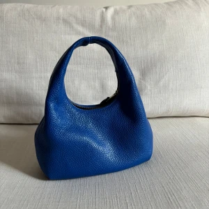 Blå handväska i skinn - Säljer en blå handväska i skinn med en dragkedja köpt från Other Stories. Väskan har en rundad form och ett bekvämt handtag. Den är i bra skick, inte använt den mycket därmed att jag säljer den. 