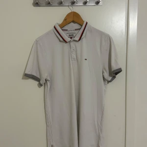 Vit pikétröja från Hilfiger Denim - Säljer en stilren vit pikétröja från Hilfiger Denim i storlek L. Tröjan har klassisk krage med röd och marinblå randdetalj samt korta ärmar. Perfekt för en avslappnad men snygg look. Passar både till vardags och fest!