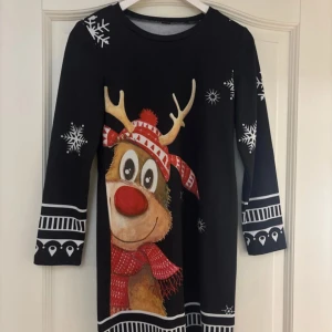 Svart julklänning med renmotiv - Superfin svart klänning med ett stort renmotiv och snöflingor, perfekt för julen! Den har trekvartsärmar och är gjord i ett mjukt material. Klänningen har en normal passform och är dekorerad med vita detaljer. Aldrig använd.