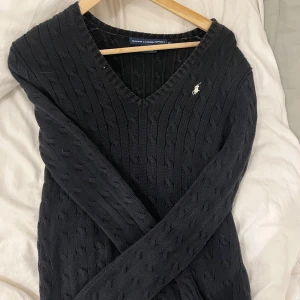 Svart stickad tröja från Ralph Lauren - Säljer en snygg svart stickad tröja från Ralph Lauren med v-ringning och deras ikoniska logga på bröstet. Perfekt för både höst och vinter, den är både stilren och bekväm. Passar till alla tillfällen, från skola till middag med vänner. 🖤