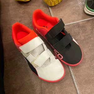 Puma fotbollsskor i svart och röd/inne skor - Säljer ett par Puma fotbollsskor i svart och röd med vita detaljer. Skorna har kardborreband för enkel på- och avtagning och en mönstrad sula för bra grepp. Perfekta för inomhusfotboll eller träning. Använda men i bra skick.