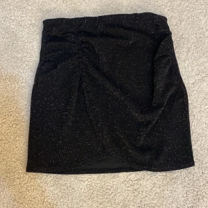 Svart glittrig kjol från Kappahl - Säljer en superfin svart glittrig kjol från Kappahl i storlek S. Kjolen har en tight passform och är perfekt för festliga tillfällen. Den har en snygg rynkdetalj framtill som ger en extra touch. Perfekt för nyår eller andra glittriga event! ✨
