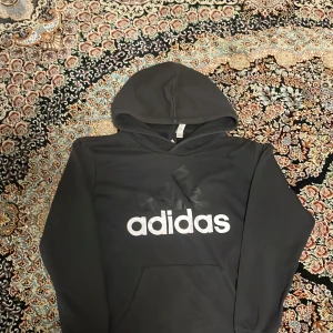 Svart hoodie från Adidas - Säljer en svart hoodie från Adidas i bra skick. Den har en stor vit Adidas-logga på framsidan och en praktisk magficka. Perfekt för höst och vinter, eller bara för att chilla i. Skön och stilren design!