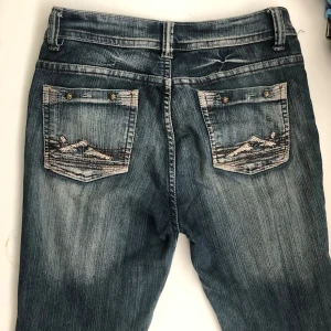 low waist bootcut y2k jeans - Säljer ett par snygga blå jeans från J.F.N med coola slitningar och broderade detaljer på bakfickorna. De har en klassisk femficksdesign och en bekväm passform. Perfekta för en avslappnad stil!
