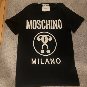 Svart t-shirt från Moschino - Säljer en svart t-shirt från Moschino med deras ikoniska logga tryckt i vitt på framsidan. T-shirten är gjord av 100% bomull och har en klassisk passform med korta ärmar. Perfekt för en stilren och trendig look!