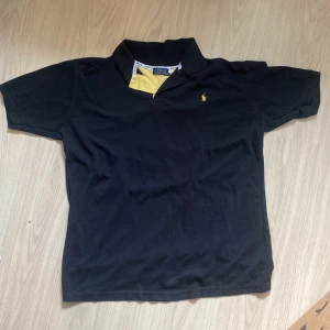 Svart Ralph lauren polo - Svart Ralph Lauren polo med gula detaljer och logga, inget tryck eller något i bak, det står XL i lappen men sitter som M