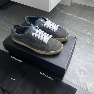 Blå sneakers från Saint Laurent - Snygga grå sneakers från Saint Laurent med vit snörning och märkets namn på sidan. Skorna har en klassisk design med en bekväm sula och är perfekta för en stilren look. Skorna är rätt så slitna därför de låga priset. För fler frågor eller bilder kom privat. Priset är ej hugget i sten!