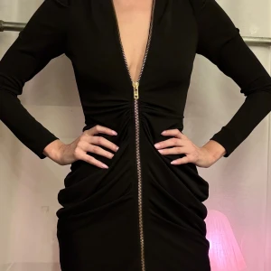 Svart klänning med dragkedja från Mary Kate & Ashley Olsen + BIKBOK - Snygg svart klänning från BIKBOK samarbete med en djup v-ringning och dragkedja framtill. Klänningen har långa ärmar och en åtsittande passform som framhäver figuren. Perfekt för en kväll ute eller en speciell tillställning.