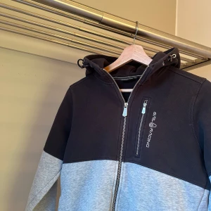 Svart och grå hoodie från Sail Racing - Snygg hoodie från Sail Racing i svart och grått med dragkedja och luva. Tröjan har en broderad logga på bröstet och huvan. Perfekt för en stilren look. Ny pris: 1500kr