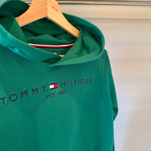 Grön hoodie från Tommy Hilfiger - Säljer en snygg grön hoodie från Tommy Hilfiger med logotyp och text 'Est. 1985' på framsidan. Perfekt för en avslappnad stil med en klassisk touch. Hoodien har en bekväm passform och är tillverkad i mjukt material. Ny pris: 1299kr