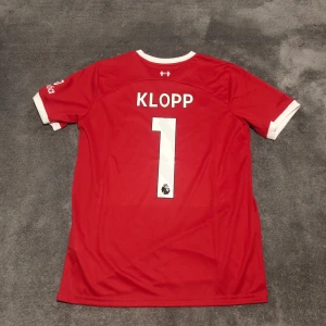 Liverpool FC | 23/24 | Klopp | Size:L | Hemmatröja | Ny, oanvänd  - Liverpools hemmatröja för säsongen 2023/2024 även sista säsongen för Liverpool legenden Jurgen Klopp. Jurgen Klopp, nr 1 på ryggen. Stadium/fan edition. Storlek L. Tröjan är en nyproducerad reprint i utmärkt kvalitet.  Skickas 1-3 dagar efter mottagen betalning. Har du frågor/funderingar är det bara att höra av dig! :)