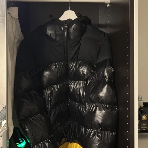 Svart pufferjacka från replay  - Snygg svart pufferjacka från Replay med glansigt material och dragkedja framtill. Jackan har en hög krage och är perfekt för kyliga dagar. Den har en modern design som passar bra till olika stilar.