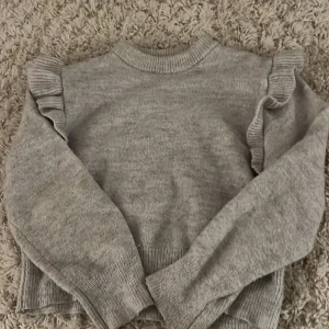 Grå tröja med volangdetaljer från Gina Tricot - Säljer en mysig grå tröja från Gina Tricot med volangdetaljer längs axlarna. Tröjan har långa ärmar och ribbade muddar vid hals, ärmslut och nederkant. Perfekt för kyliga dagar!