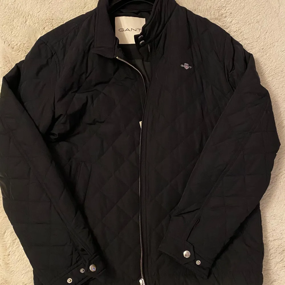 Snygg svart quiltad jacka från Gant med dragkedja och tryckknappar vid kragen. Jackan har en stilren design med långa ärmar och diskret logga på bröstet. Storlek M. Helt ny jacka. Nypris 2000kr. Takit.