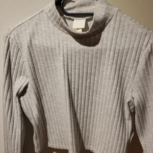 Ribbad polotröja i grått och vitt - Säljer två ribbade (croppad) polotröjor i grått och vitt. Tröjorna har en stilren design med långa ärmar och en hög krage. Perfekta för en stilren look under kyligare dagar. 