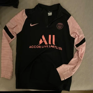 Svart och rosa fotbollströja från Nike - Snygg svart fotbollströja med rosa detaljer från Nike. Tröjan har långa ärmar i rosa med svarta ränder och en halv dragkedja framtill. PSG-logga och sponsortryck på bröstet samt 'Visit Rwanda' på ryggen.