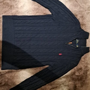 Mörkblå stickad tröja från Ralph Lauren - Säljer en snygg mörkblå stickad tröja från Ralph Lauren med dragkedja och röd broderad logga på bröstet. Tröjan har ett klassiskt kabelstickat mönster och långa ärmar. Perfekt för kyligare dagar! Original pris 2695