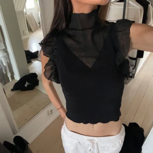 Svart topp med volangdetaljer från Zara - Snygg svart topp från Zara med volangdetaljer och hög krage. Toppen är ärmlös och har en elegant design som passar perfekt för en stilren look. Tillverkad i ett lätt och luftigt material.