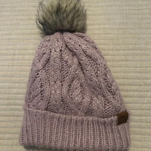 Stickad mössa med pälsboll från H&M - Säljer en stickad mössa från H&M med en fluffig pälsboll på toppen. Mössan har ett snyggt flätmönster och finns i både lila och grått. Perfekt för kalla dagar! 50 kr för båda.