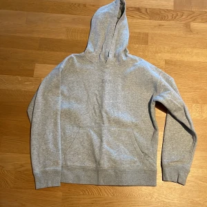 Grå hoodie från GAP Kids - Säljer en klassisk grå hoodie från GAP Kids i storlek XXL (14-16). Tröjan är tillverkad av 77% bomull och 23% återvunnen polyester. Den har en praktisk känguruficka och en bekväm huva. Perfekt för en avslappnad stil.