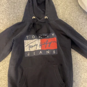 hoodie från Tommy Hilfiger - Säljer en hoodie från Tommy Hilfiger med stor logga på bröstet i blått, vitt och rött. Den har en klassisk känguruficka och justerbar huva med snören. Perfekt för en avslappnad stil.