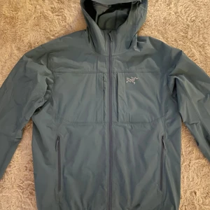 Arc'teryx vindjacka - Säljer en blå vindjacka från Arc'teryx med huva och dragkedja, storlek Medium. Jackan har en stilren design med två sidofickor och två bröstfickor, perfekt för utomhusaktiviteter. Märkets logga syns på bröstet. Nypris 300€.