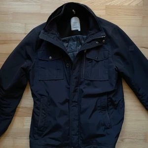 Mörkblå fieldjacket från J.Lindeberg - Snygg svart jacka från J.Lindeberg med dragkedja och knappar. Använd fåtal gånger 8/10 cond, pris 1149kr!