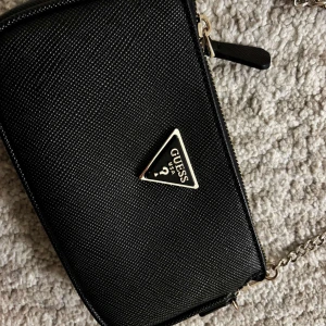 Svart axelväska från Guess - Snygg svart axelväska från Guess med en elegant kedjerem och det ikoniska Guess-emblemet på framsidan. Perfekt för att bära dina nödvändigheter med stil. Väskan har en praktisk dragkedja för säker förvaring.