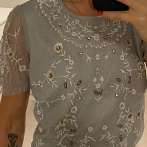 Ljusblå blus med broderade blommor - Säljer en ljusblå blus med korta ärmar och vackra broderade blommor. Blusen har en öppen rygg med snörning och små pärlor som detaljer. Perfekt för en somrig look! Stlk L men passar S och M