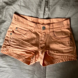 Persikofärgade shorts från KappAhl - Snygga persikofärgade shorts från KappAhl med klassisk femficksdesign och knappgylf. Perfekta för varma sommardagar. De har en broderad detalj på linningen och lågmidjade.
